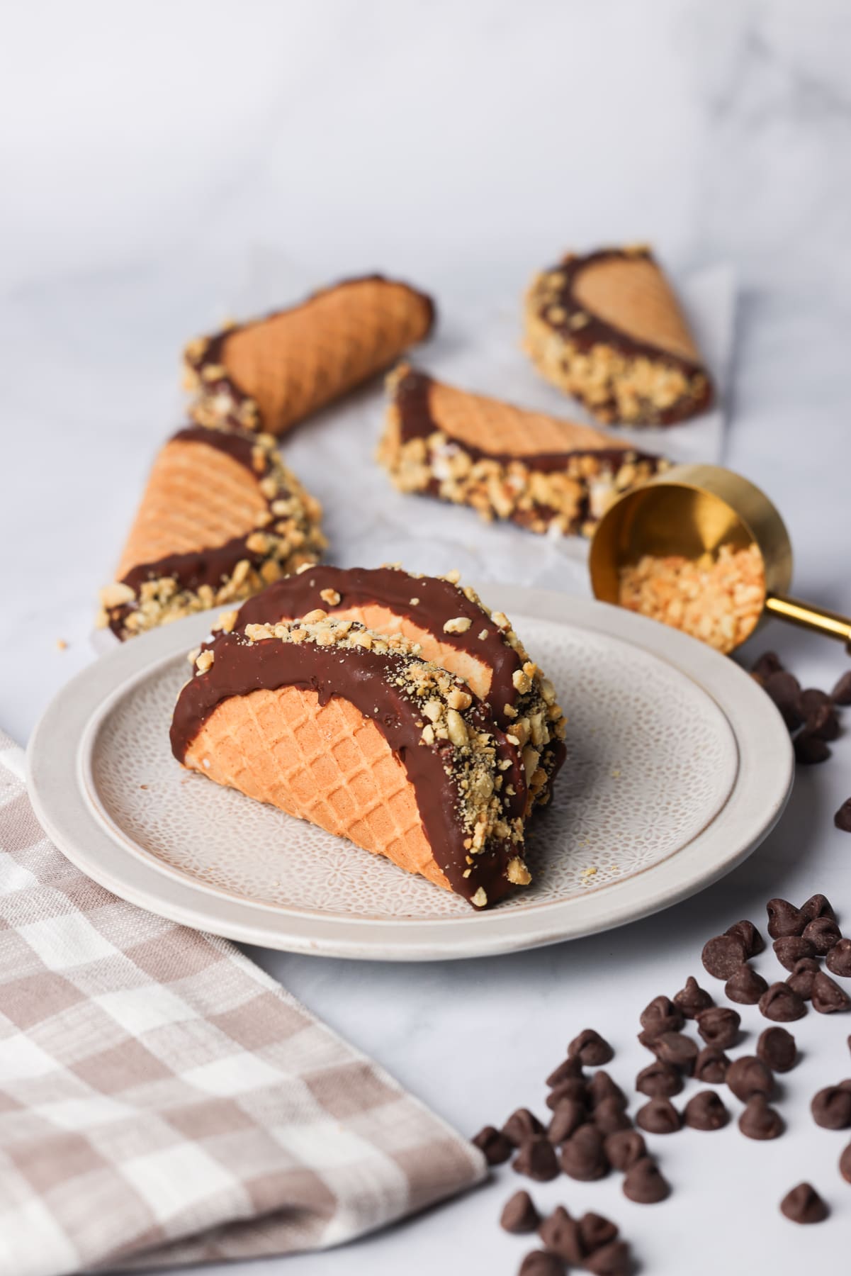 choco tacos that are filled with ice cream. they are on a plate. there are chocolate chips and a measuring cup filled with chopped nuts.