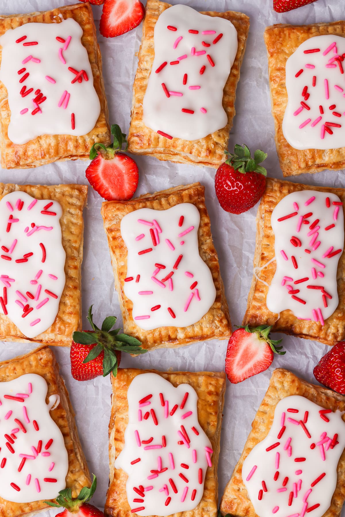 9 pop tarts with pink and red sprinkles. There are fresh strawberries surrounding the pop tarts.